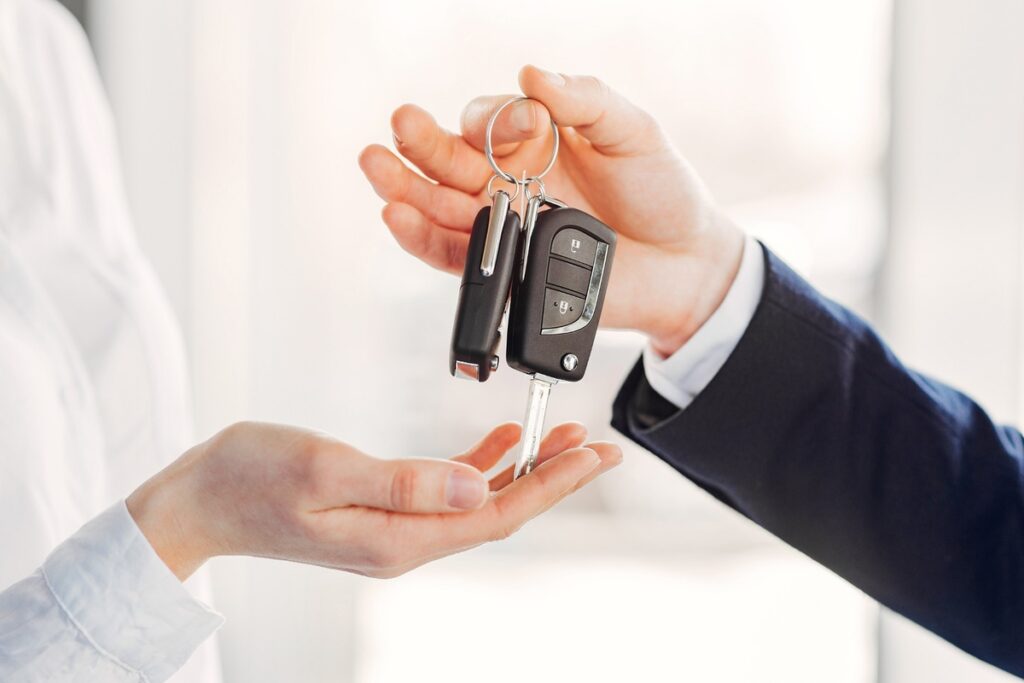 Mão de uma pessoa entregando a chave de um carro para outra, simbolizando a compra, aluguel ou entrega de veículo. O cenário é claro e moderno, e as mãos pertencem a pessoas vestidas formalmente, uma delas com terno e outra com camisa branca.